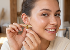 A close-up lifestyle shot of a woman wearing a gold and white bee stud earring, holding the earring in her fingers to show its details against a warm, softly blurred indoor background.